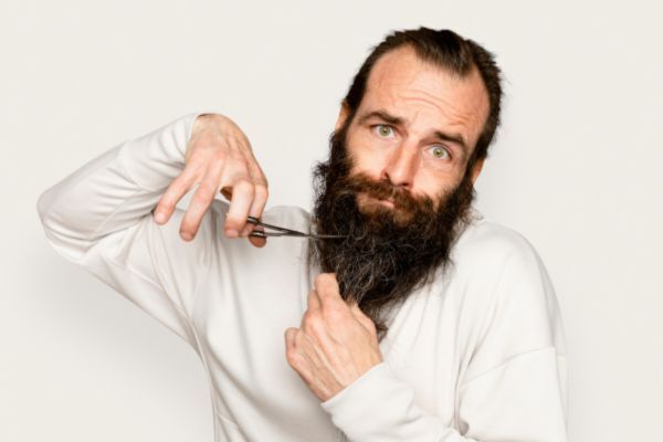A man with a long, wild beard and an expressive face awkwardly attempts to cut his beard with a pair of scissors. His uncertain expression and body language add humor to the grooming attempt.