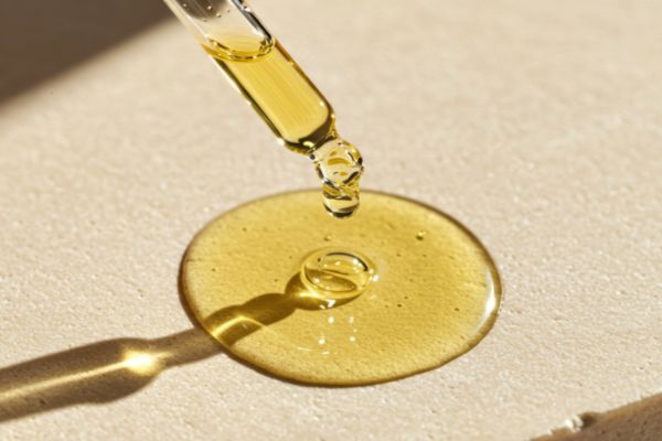 A close-up of a glass dropper releasing golden beard oil onto a smooth beige surface, highlighting the oil’s rich texture and moisturizing quality.