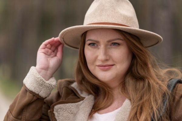 Smiling woman with long, wavy light brown hair wearing a tan felt hat and brown shearling jacket, standing outdoors with a blurred natural background.