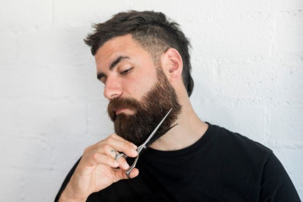 A man with a full beard carefully trims the bottom edge with a pair of scissors, concentrating as he shapes it evenly. He wears a black shirt and stands against a white wall.