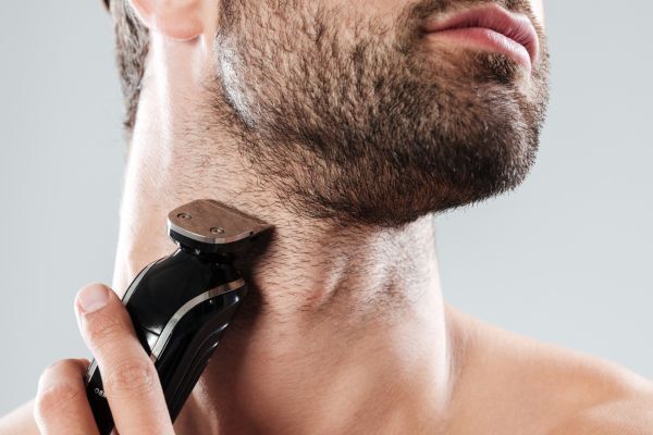 A close-up of a man using an electric trimmer to shape the neckline of his beard. His skin is slightly stretched as he guides the trimmer along his neck for a clean edge.