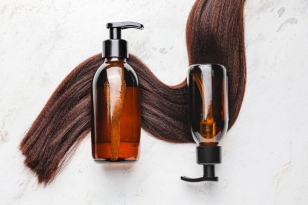 Two amber pump bottles placed over long, healthy brown hair strands on a light marble surface, representing hair growth shampoos and conditioners.
