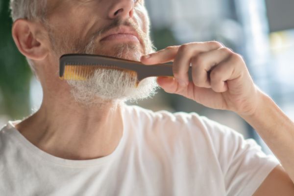A close-up of a man gently combing his gray beard using a small grooming comb. He wears a plain white t-shirt, with sunlight casting a soft glow on his beard and hand.