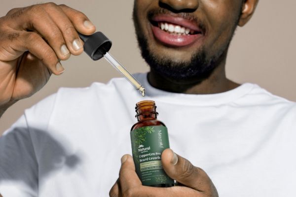 A smiling man in a white shirt holds a dropper bottle of beard oil labeled “Natural CopperGro Pro Beard Growth” and is about to apply the oil using the dropper.