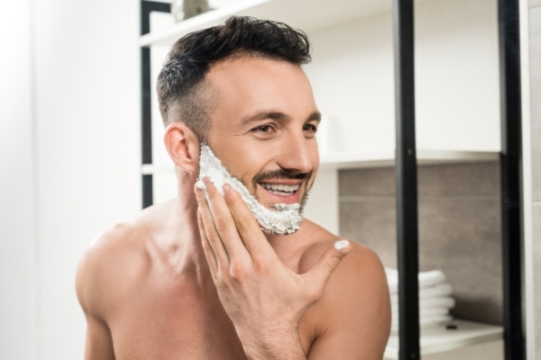 A smiling man with a trimmed beard applies shaving cream to his cheek while standing in a modern bathroom. His expression and grooming suggest a self-care routine focused on facial hair maintenance.