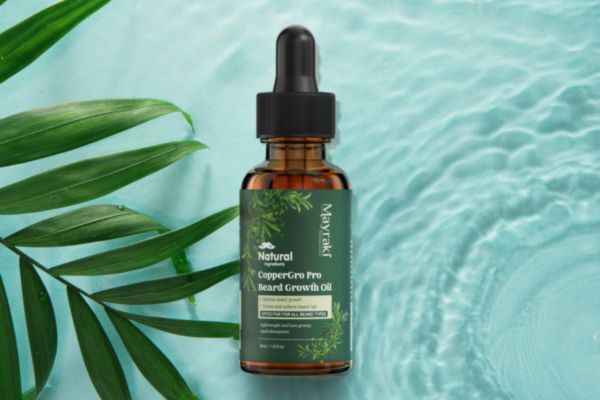 A dropper bottle of Mayrack Natural CopperGro Pro Beard Growth Oil is displayed on a light blue water ripple background with green palm leaves. The label highlights natural ingredients and beard growth benefits.