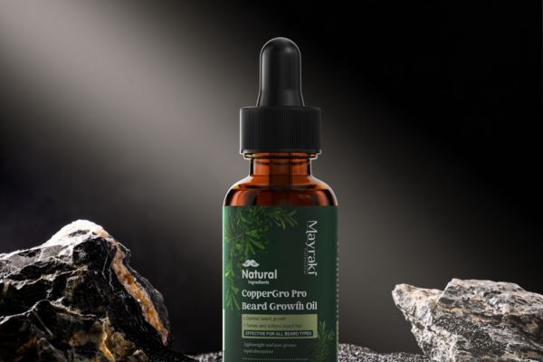 A bottle of Mayraki Natural CopperGro Pro Beard Growth Oil stands upright between dark, textured rocks under a spotlight. The green label highlights that the product is natural, promotes beard growth, and is suitable for all beard types.