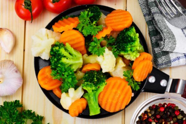 A colorful mix of steamed vegetables including broccoli, cauliflower, and crinkle-cut carrots, garnished with fresh parsley in a black skillet on a wooden surface. Surrounding elements like garlic, tomatoes, and peppercorns add a fresh, healthy food vibe.