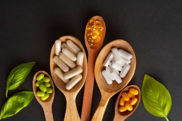 A variety of vitamins and supplements, including capsules and softgels in different colors, are displayed on wooden spoons surrounded by fresh green leaves, suggesting natural support for hair and overall wellness.