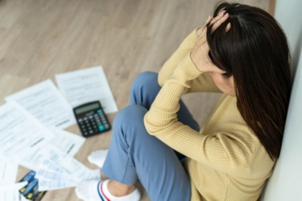 This image shows a distressed woman sitting on the floor with her head in her hands, surrounded by scattered bills, a calculator, and credit cards. The scene conveys financial stress or anxiety, likely due to overwhelming debt or budgeting challenges.