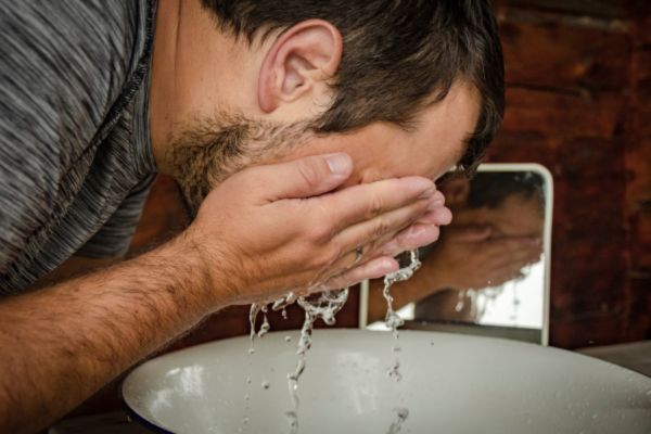 A man with a short beard splashes water on his face over a white basin, captured mid-routine in a rustic bathroom setting with a small mirror in the background. This image conveys a cleansing or grooming process, possibly related to beard care.