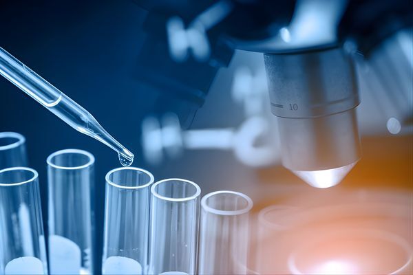 A close-up of lab equipment with a pipette releasing liquid into test tubes under a microscope&rsquo;s lens. The scientific setting suggests research and formulation of vitamins and supplements related to hair growth effectiveness.