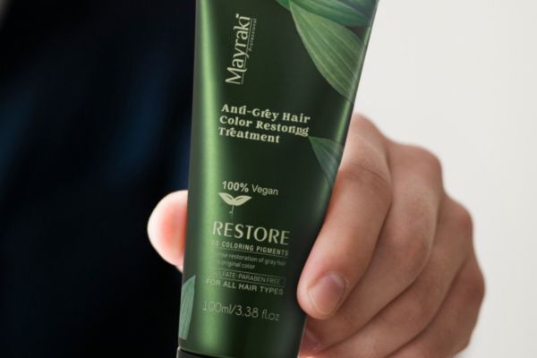 A hand holds a green tube of Mayraki Anti-Grey Hair Color Restoring Treatment labeled as 100% vegan and free from sulfates and parabens. The product highlights the benefits of natural anti-grey treatments that aim to reverse greying without harsh chemicals.