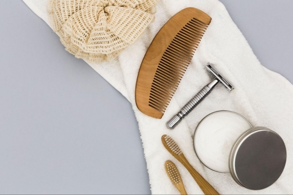 A flat lay of men's grooming essentials on a white towel, including a wooden comb, safety razor, round tin of shaving cream, two bamboo toothbrushes, and a textured bath pouf, arranged neatly against a gray background.