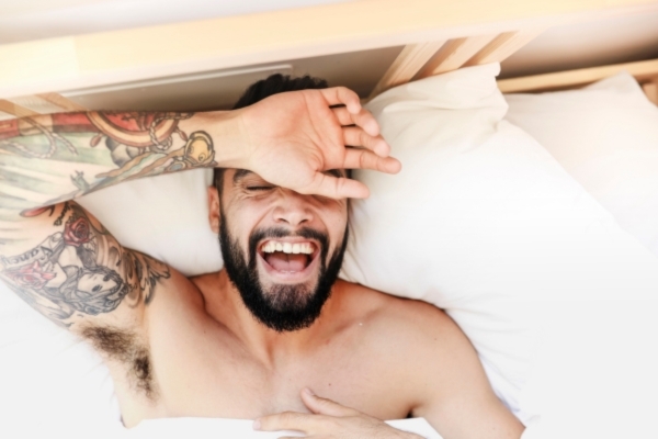 A bearded man with tattoos stretches and laughs while lying shirtless in bed, with white pillows and sunlight softly lighting the scene. The image conveys a relaxed, confident vibe, possibly suggesting a refreshed look tied to grooming or self-care.