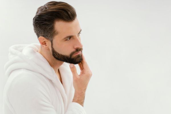 A man in a white bathrobe touches his neatly groomed beard with a contemplative look, standing against a clean, white background. His pose reflects care and confidence in his grooming routine.
