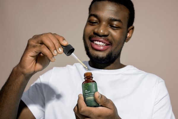 A smiling man in a white shirt holds an open bottle of Mayraki CopperGro Pro Beard Growth Oil, using the dropper to apply the product. His cheerful expression and well-groomed beard highlight the satisfaction and care associated with using natural beard grooming products.